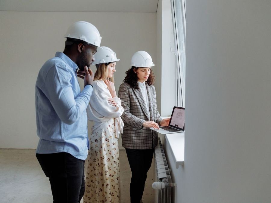 Three colleagues in white hard hats reviewing plans on a laptop inside a building under renovation