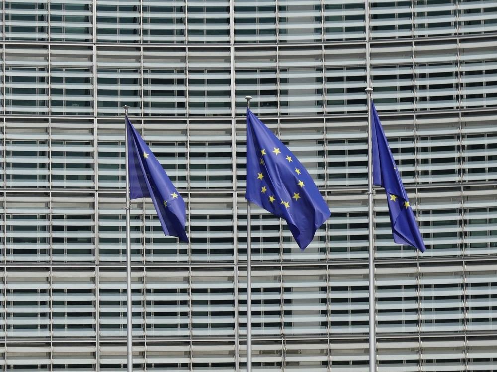Three European Union flags on flagpoles in front of a glass-fronted office building
