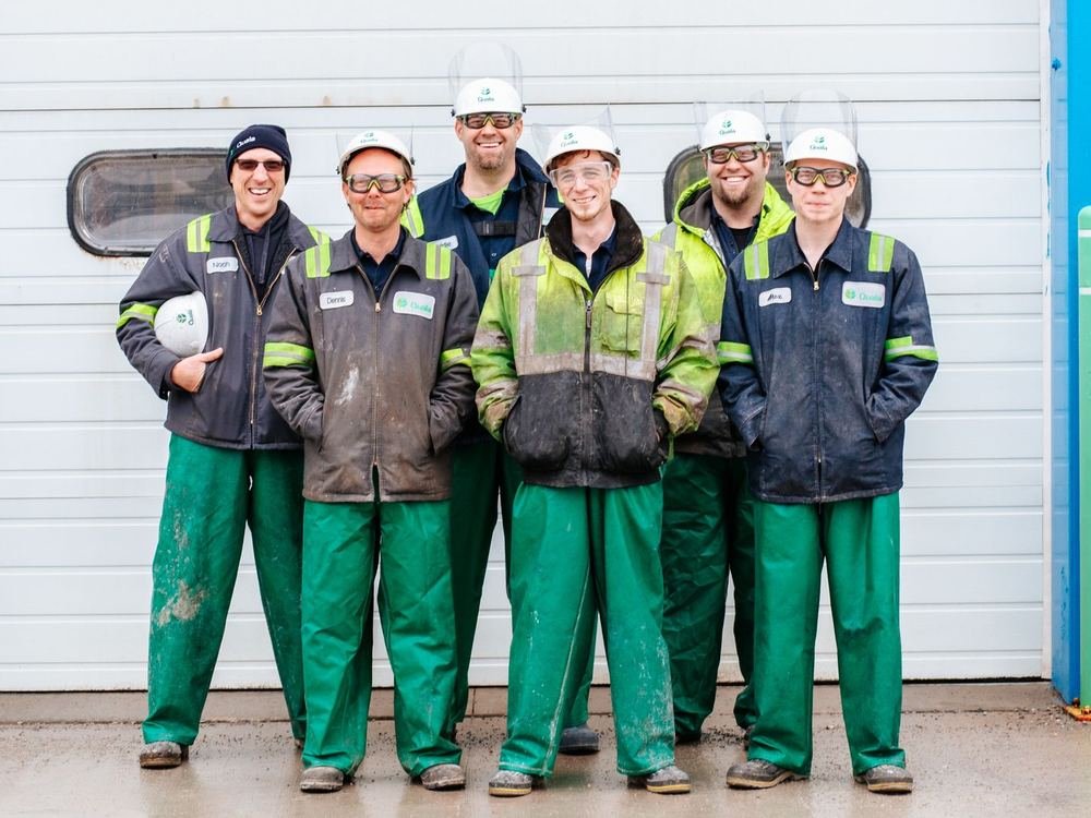 A team of six workers in hard hats, safety glasses and work jackets smiling together outside an industrial building